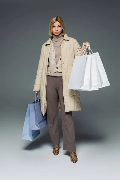 Full length of blonde young woman in winter puffer coat holding shopping bags on grey — Stock Photo
