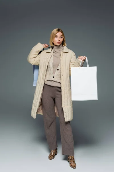 Mujer de moda en chaqueta de invierno y cuello alto sosteniendo bolsas de compras sobre fondo gris - foto de stock