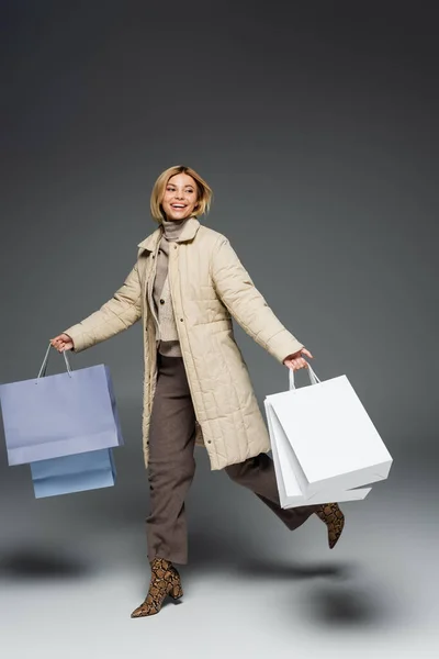 Весела жінка в зимовій куртці тримає сумки для покупок під час роботи на сірому фоні — стокове фото