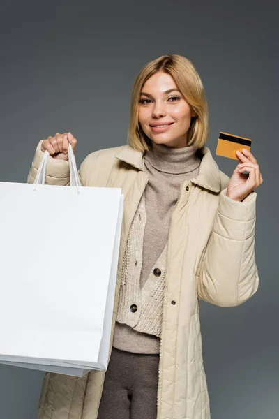 Joven sonriente en traje de invierno sosteniendo bolsas de compras y tarjeta de crédito aislada en gris - foto de stock