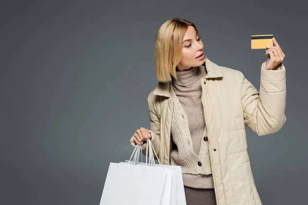 Jeune femme en tenue chaude regardant la carte de crédit tout en tenant des sacs à provisions isolés sur gris — Photo de stock