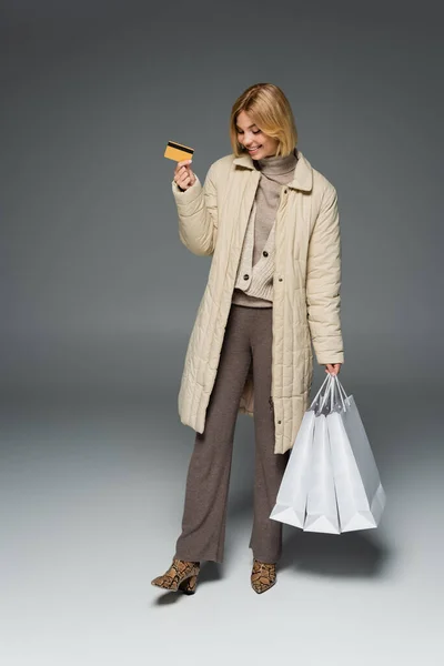 Smiling woman in winter outfit holding credit card and shopping bags on grey background — Stock Photo