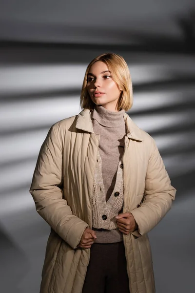 Модная молодая женщина в бежевом зимнем наряде смотрит в сторону на абстрактном сером фоне — стоковое фото