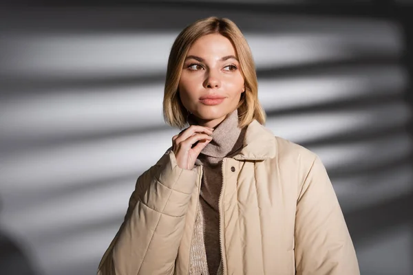 Hübsche junge Frau in beiger Winterjacke schaut vor abstrakt grauem Hintergrund weg — Stockfoto