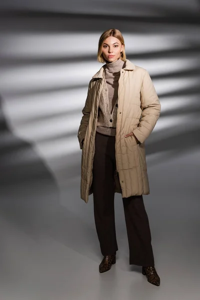 Jeune femme branchée en tenue d'hiver posant et regardant la caméra sur fond gris abstrait — Photo de stock