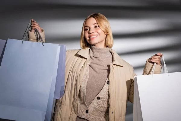 Mulher loira feliz no inverno jaqueta segurando compras no fundo cinza abstrato — Fotografia de Stock