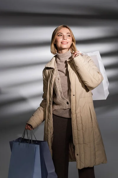 Mujer alegre en chaqueta de invierno sosteniendo bolsas de compras sobre fondo gris abstracto - foto de stock