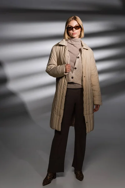Полная длина модной женщины в зимней куртке и солнцезащитных очках, стоящих на абстрактном сером фоне — стоковое фото