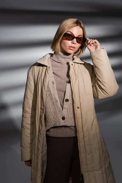 Trendy junge Frau in Sonnenbrille und Winterjacke posiert auf abstrakt grauem Hintergrund — Stockfoto