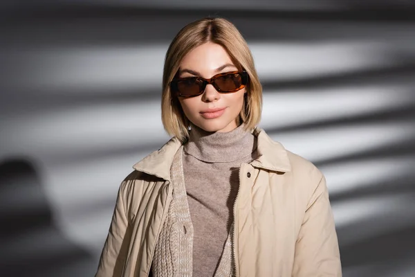 Blonde Frau mit Sonnenbrille und Winterjacke auf abstrakt grauem Hintergrund — Stockfoto