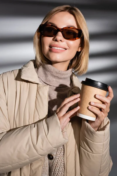 Donna sorridente in occhiali da sole e giacca invernale in possesso di caffè per andare su sfondo grigio astratto — Foto stock