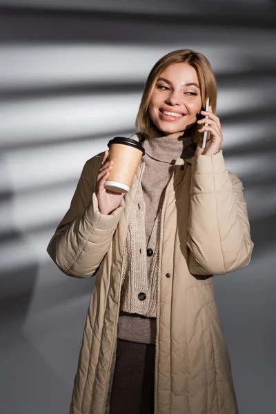 Jovem alegre no casaco de inverno segurando bebida takeaway e falando no smartphone em fundo cinza abstrato — Fotografia de Stock