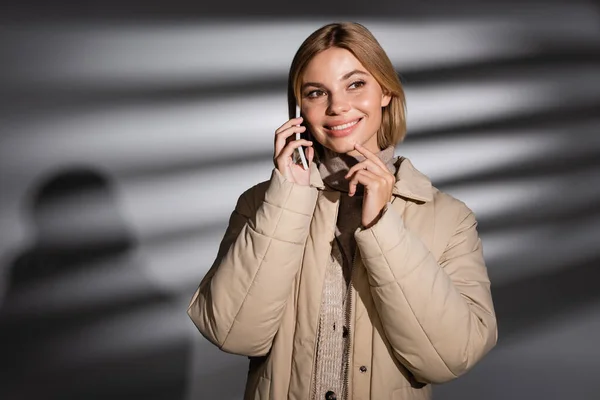 Mujer sonriente en chaqueta de invierno beige hablando en smartphone sobre fondo gris abstracto - foto de stock