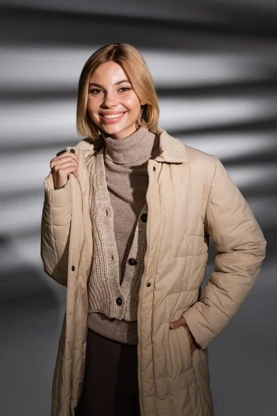 Cheerful young woman in winter jacket looking at camera on abstract grey background — Stock Photo