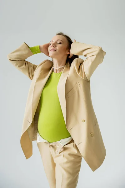 Mujer embarazada de moda en chaqueta beige tocando el cabello aislado en gris - foto de stock