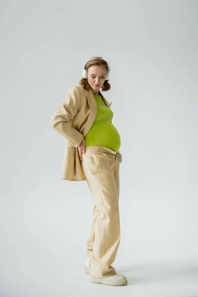 Jeune femme enceinte en costume beige et écouteurs debout sur fond gris — Photo de stock