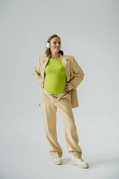 Веселая беременная женщина в костюме слушает музыку в наушниках на сером фоне — стоковое фото
