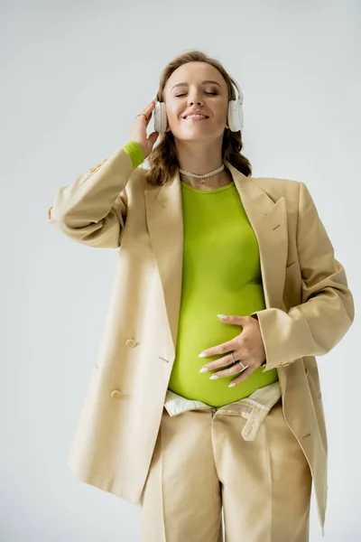 Щаслива вагітна жінка в куртці торкається живота і слухає музику в навушниках ізольовано на сірому — стокове фото