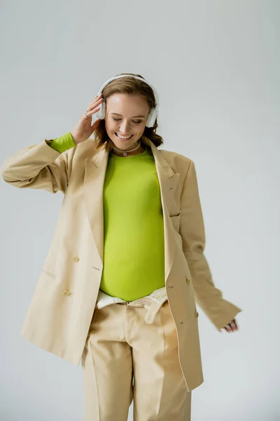 Fashionable pregnant woman smiling while listening music in headphones isolated on grey — Stock Photo