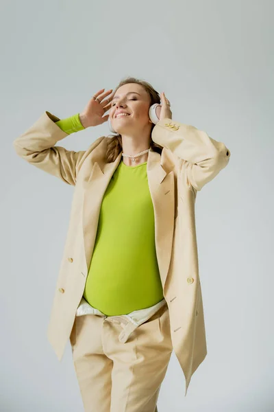 Glücklich schwangere Frau im Anzug hört Musik in Kopfhörern isoliert auf grau — Stockfoto