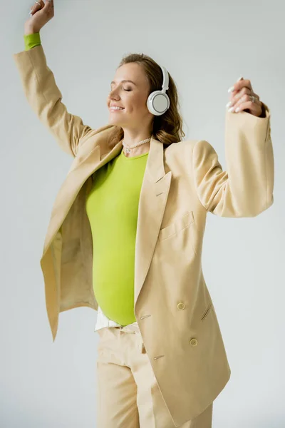 Cheerful pregnant woman in headphones and jacket standing isolated on grey — Stock Photo