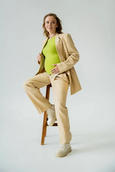 Mujer embarazada de moda en blusa y traje sentado en silla sobre fondo gris - foto de stock