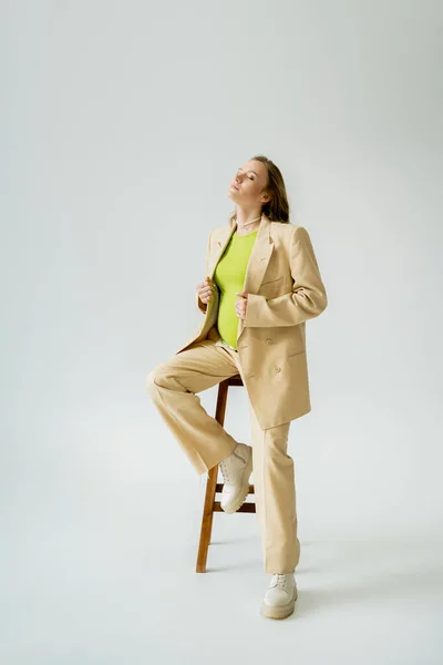 Модная беременная женщина в костюме позирует возле стула на сером фоне — стоковое фото