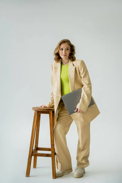 Longitud completa de la mujer embarazada de moda sosteniendo el ordenador portátil y mirando hacia otro lado cerca de la silla sobre fondo gris - foto de stock