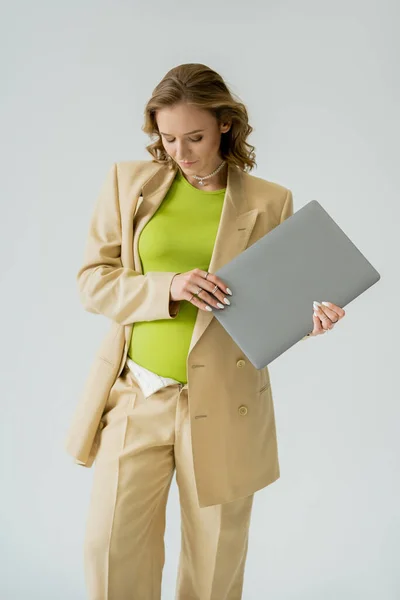 Стильная беременная фрилансер в бежевом костюме, держащая ноутбук изолированным на сером — стоковое фото
