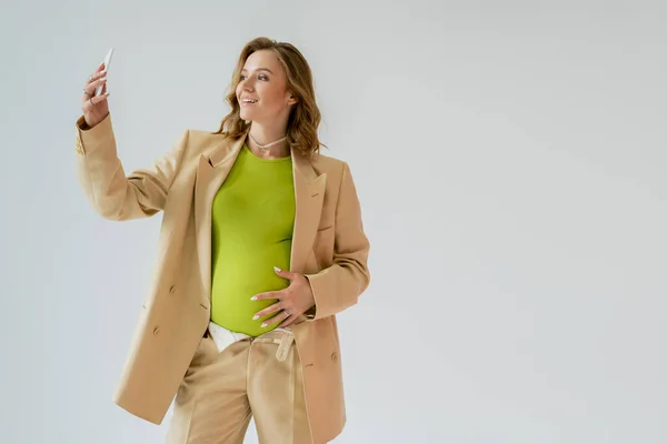 Donna incinta alla moda in giacca e cravatta scattare selfie sul cellulare isolato su grigio — Foto stock