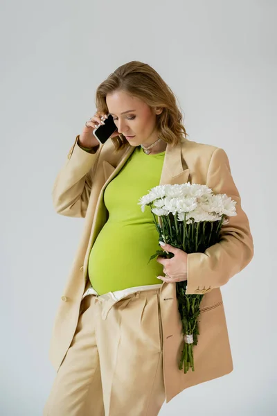 Jeune femme enceinte en costume parlant sur smartphone et tenant des fleurs isolées sur gris — Photo de stock