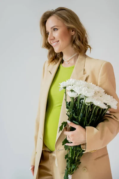 Fröhliche Schwangere im Anzug hält Blumenstrauß in der Hand und schaut vereinzelt auf grau — Stockfoto
