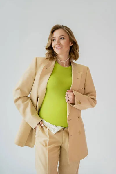 Mujer embarazada sonriente posando en traje beige aislado en gris - foto de stock