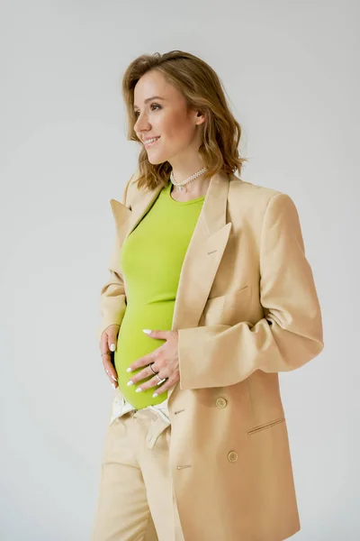 Стильная беременная женщина в куртке, касающаяся живота, изолированного от серого — стоковое фото