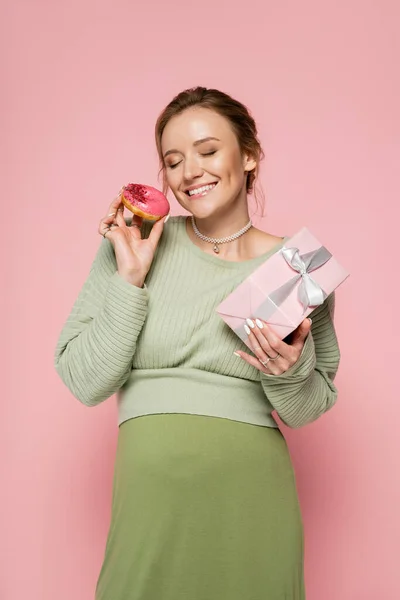 Улыбающаяся беременная женщина в свитере держит пончик и подарок на розовом фоне — стоковое фото