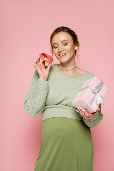 Cheerful pregnant woman holding present and donut on pink background — Stock Photo
