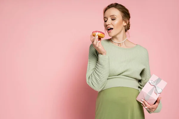 Молодая беременная женщина с подарочной коробкой и вкусным пончиком на розовом фоне — стоковое фото