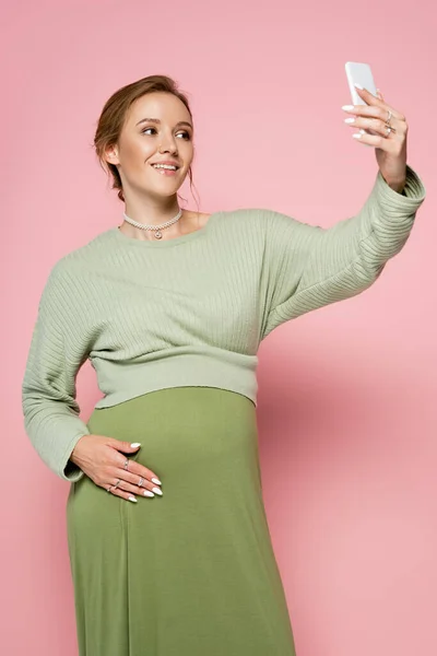 Положительная беременная женщина делает селфи на смартфоне на розовом фоне — стоковое фото
