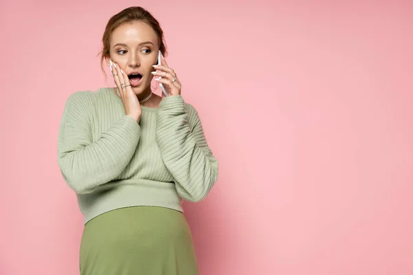 Shocked pregnant woman in green outfit talking on cellphone on pink background — Stock Photo