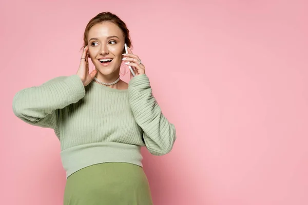 Mujer embarazada positiva en traje verde hablando en el teléfono inteligente sobre fondo rosa - foto de stock