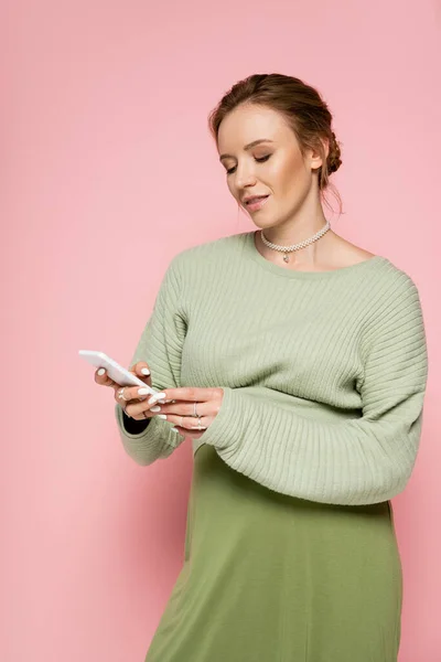 Stylish pregnant woman in green outfit using mobile phone on pink background — Stock Photo