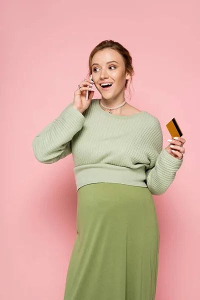 Mujer embarazada emocionada en traje verde con tarjeta de crédito y hablando en el teléfono inteligente sobre fondo rosa - foto de stock