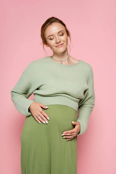 Mujer embarazada con estilo en suéter tocando vientre aislado en rosa - foto de stock