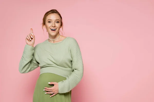 Eccitato donna incinta in abito verde che punta con il dito isolato su rosa — Foto stock