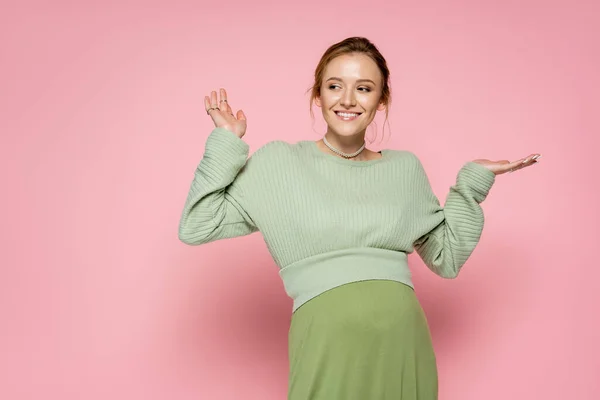 Femme enceinte positive en tenue verte tendance pointant avec les mains sur fond rose — Photo de stock