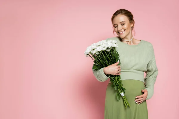 Femme enceinte positive en tenue verte regardant bouquet sur fond rose — Photo de stock