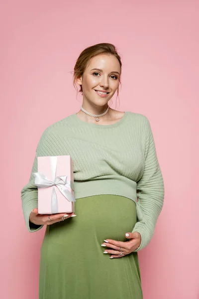 Fröhliche Schwangere in grünem Outfit hält Geschenk in der Hand und blickt vereinzelt in die Kamera auf rosa — Stockfoto