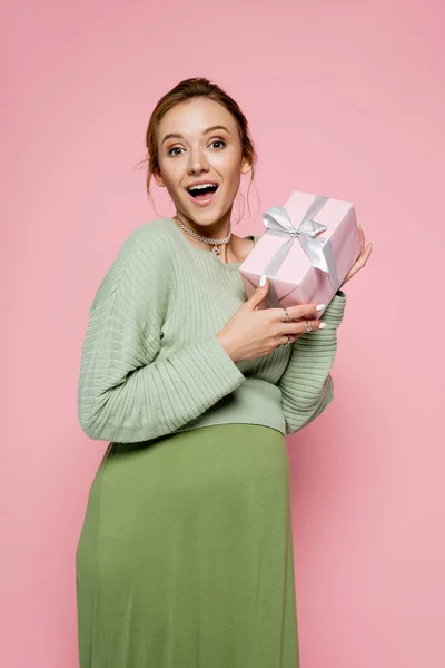 Возбужденная беременная женщина в зеленом костюме держит подарок изолирован на розовый — стоковое фото