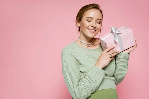 Positive pregnant woman holding present with bow isolated on pink — Stock Photo