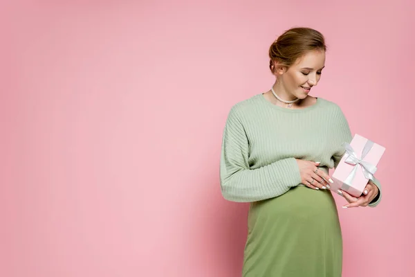 Happy pregnant woman in green outfit holding present on pink background — Stock Photo